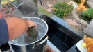 Outdoor cooking scene with a hand placing a lobster into a steaming pot on a built-in stainless steel burner, garden and pond backdrop