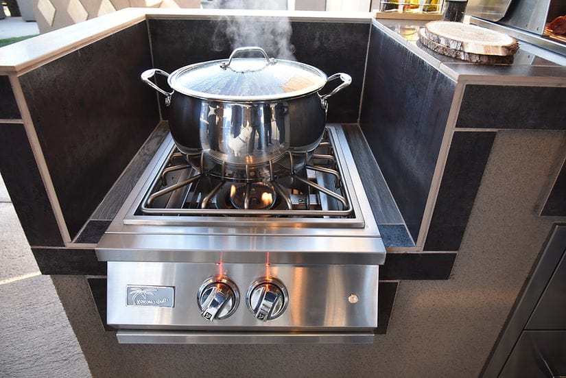 Outdoor stainless steel power burner in a built-in setup, cooking a steaming pot, with illuminated control knobs and a modern backsplash