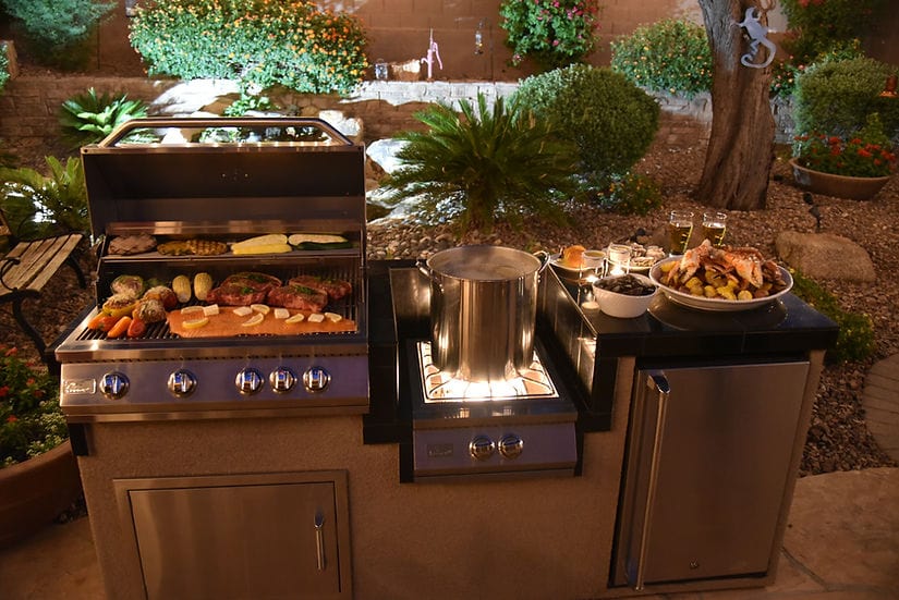Outdoor kitchen setup with a grill loaded with food, a boiling pot on a burner, and a side counter with prepared dishes, set in a garden