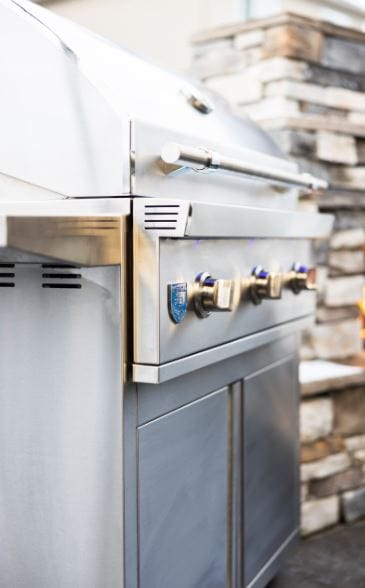 Side view of a stainless steel outdoor grill with closed lid, illuminated control knobs, and stone fireplace in the background.
