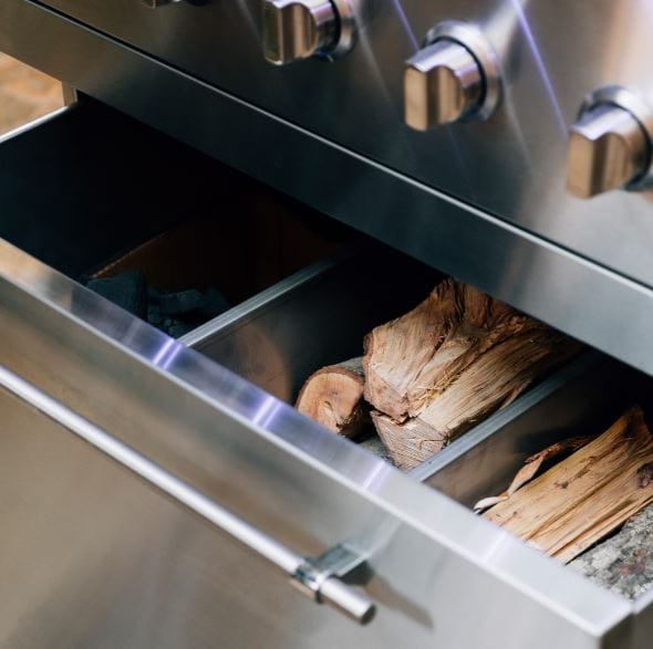 Close-up of a stainless steel grill drawer containing wood logs and charcoal, designed for versatile outdoor cooking options.