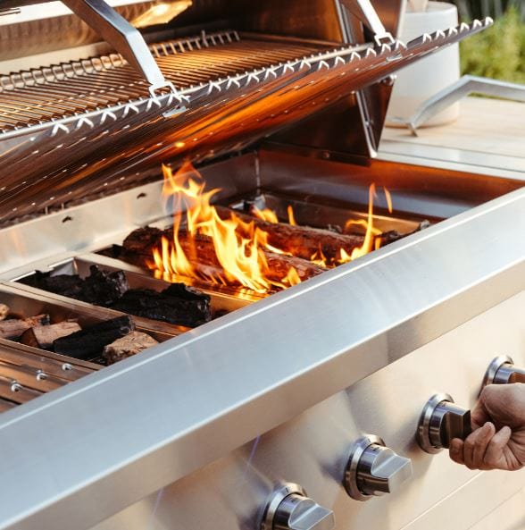 Open stainless steel grill with flames and charcoal, adjustable control knobs, and a warming rack, designed for outdoor cooking.
