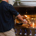 Person grilling meat over open flames on a stainless steel grill with illuminated knobs, bread and a skillet visible on the warming rack.
