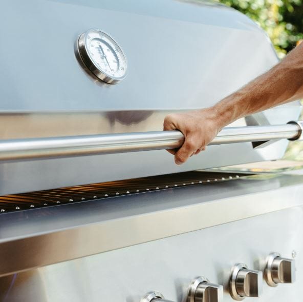 Close-up of a person opening a stainless steel grill lid with a built-in thermometer and sleek handle, showcasing modern design.