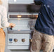 Two people standing near an open stainless steel grill with interior lights, warming rack, and control knobs, ready for outdoor cooking.