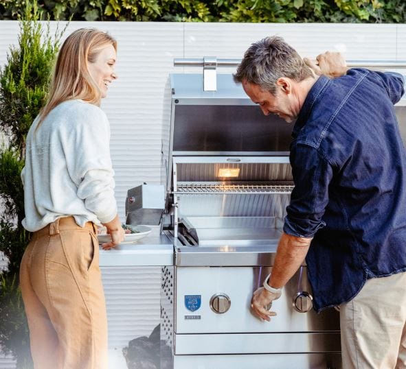 Man and woman preparing food at an open stainless steel grill in an outdoor setting with greenery, featuring interior lights and knobs.