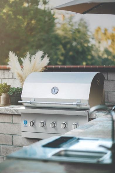 Built-in stainless steel grill with a closed lid, thermometer, and outdoor kitchen setup surrounded by greenery and modern decor.