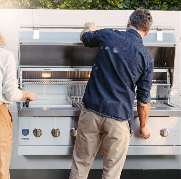 Two people using a large stainless steel grill with open lids, interior lighting, warming racks, and spacious cooking grates in a backyard.