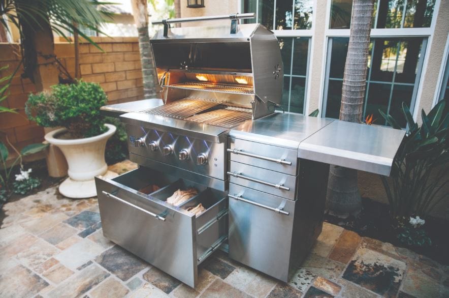 Outdoor stainless steel grill with open lid, illuminated interior, LED-lit knobs, wood storage drawer, side shelves, and a patio setting.