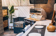 Modern outdoor kitchen with a stainless steel grill, side burner, dining setup, potted plant, and stylish black light fixture.