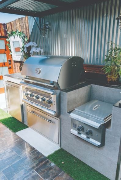 Modern outdoor kitchen with a built-in stainless steel grill, side burner, storage cabinets, and corrugated metal siding decor.