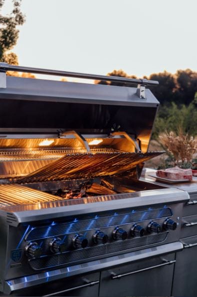 Stainless steel grill with open lid, interior lights, raised warming rack, and illuminated control knobs in a serene outdoor setting.