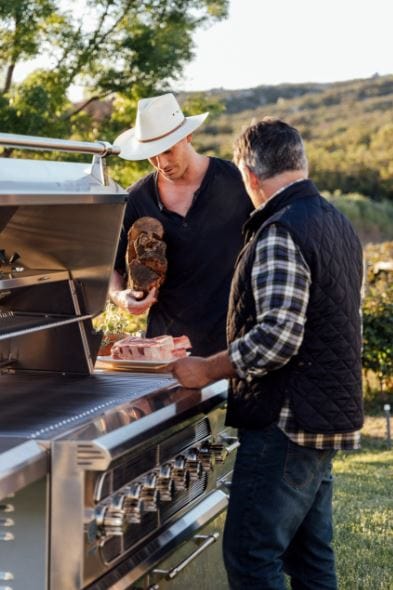 Two people preparing food on a stainless steel grill in a scenic outdoor setting, surrounded by greenery and a relaxed atmosphere.