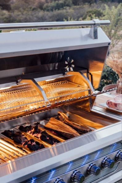 Open stainless steel grill with wood logs burning, illuminated cooking grates, warming rack, and control knobs in an outdoor setting.