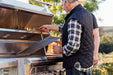 Two people working with an open stainless steel grill, adjusting the cooking grates in a scenic outdoor setting with greenery.