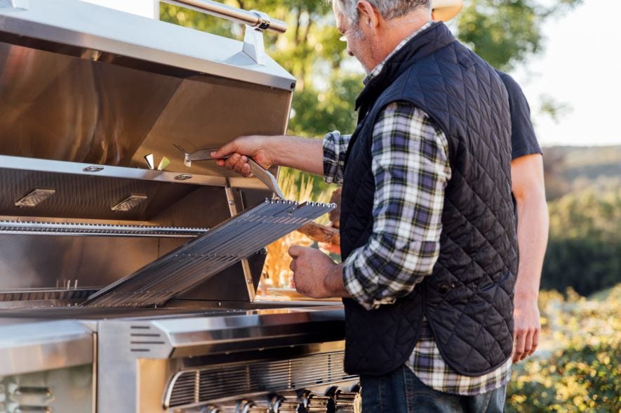 Two people working with an open stainless steel grill, adjusting the cooking grates in a scenic outdoor setting with greenery.