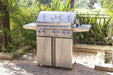 Stainless steel freestanding propane BBQ grill with side shelves on a stone patio surrounded by lush greenery