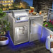 Modern outdoor kitchen setup with stainless steel grill, refrigerator, drawers, drinks, and food on counter, with blue LED lighting.
