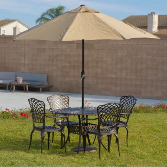 Outdoor dining set with a round table, four lattice-back chairs, and a beige umbrella in a sunny backyard lawn setting with red flowers.