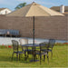 Outdoor dining set with a round table, four lattice-back chairs, and a beige umbrella in a sunny backyard lawn setting with red flowers.