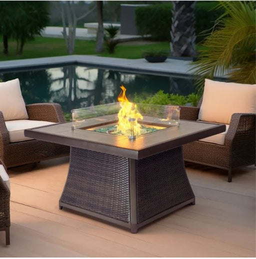 A square fire pit table with a glass enclosure, surrounded by cushioned chairs on a wooden deck, with a pool in the background.