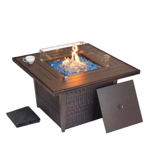 Square outdoor fire pit table with glass wind guard, blue fire glass, and a removable panel, set on a woven wicker base with a cup nearby.