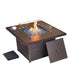 Square outdoor fire pit table with glass wind guard, blue fire glass, and a removable panel, set on a woven wicker base with a cup nearby.