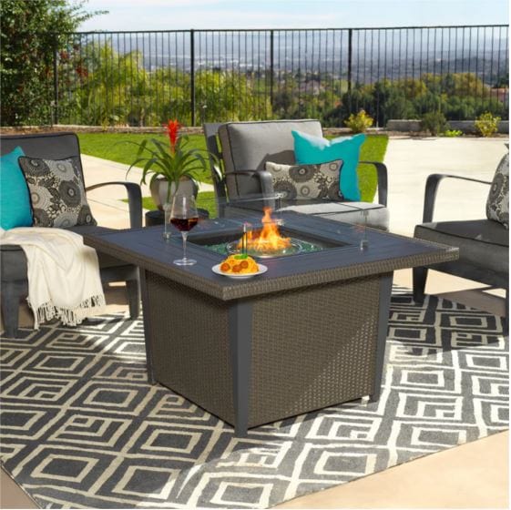 Outdoor fire pit table on a patterned rug, surrounded by patio chairs with colorful cushions, set against a scenic backyard view.