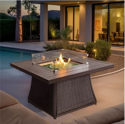 Modern outdoor fire pit table with wicker base and tempered glass wind guard, set on a patio near a pool, creating a cozy ambiance.