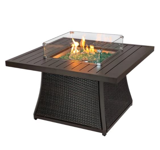 Square outdoor fire pit table with a slatted tabletop, wicker base, glass wind guard, and decorative fire glass, showcasing a lit flame.