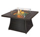 Square outdoor fire pit table with a slatted tabletop, wicker base, glass wind guard, and decorative fire glass, showcasing a lit flame.