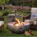 Modern outdoor fire pit with a concrete finish, surrounded by wicker chairs, wine glasses, and cozy decor on a lush green lawn.