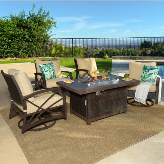 Outdoor patio setup featuring a rectangular fire pit table with flames, four tan cushioned swivel chairs, and a scenic backyard view.