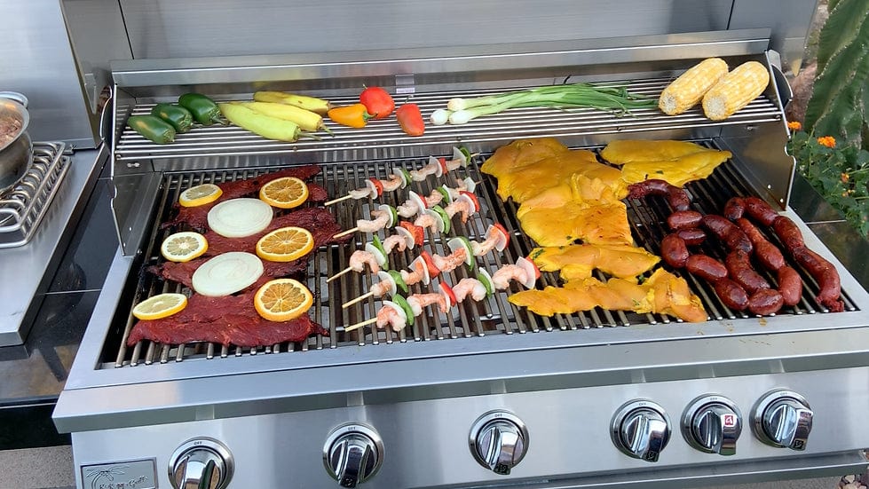 Stainless steel gas grill with a variety of foods cooking, including meats, vegetables, shrimp skewers, corn, and citrus garnishes
