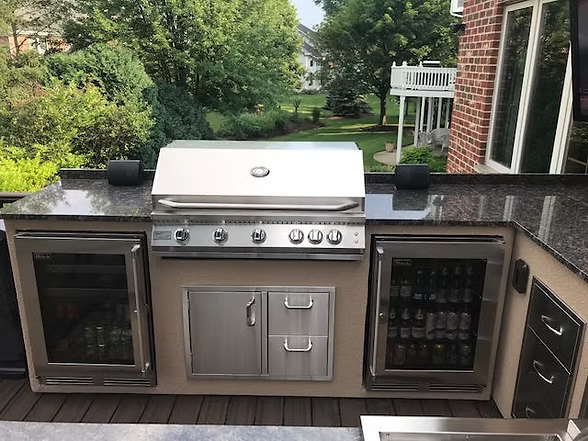 Outdoor kitchen setup featuring a stainless steel built-in gas grill, dual beverage refrigerators, storage cabinets, and granite countertops overlooking a backyard.