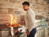 Man grilling steak on a built-in stainless steel burner with red-lit knobs, open flame visible, in an outdoor kitchen with stone decor.