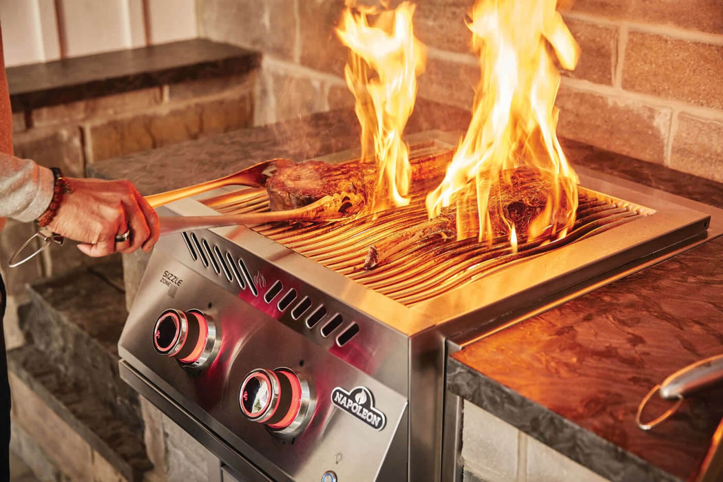 Close-up of steaks grilling over open flames on a built-in stainless steel burner with red-lit knobs in an outdoor kitchen setup.