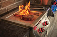 Steak grilling over open flames on a built-in stainless steel burner with red-lit knobs, part of an outdoor kitchen with stone accents.