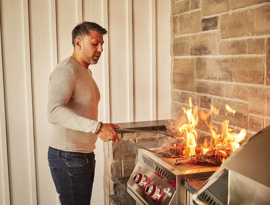 Man grilling meat over open flames on a stainless steel built-in burner with red-lit knobs, in an outdoor kitchen with stone accents.