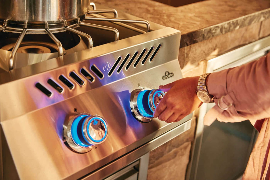 Person adjusting blue-lit control knobs on a stainless steel outdoor burner with sturdy pot supports, set in a stone-accented kitchen.