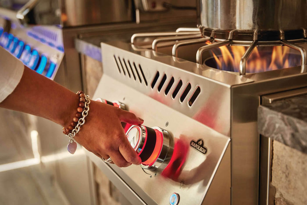 Person adjusting red-lit control knobs on a stainless steel outdoor burner with a pot cooking over a visible flame, in a stone-accented kitchen.