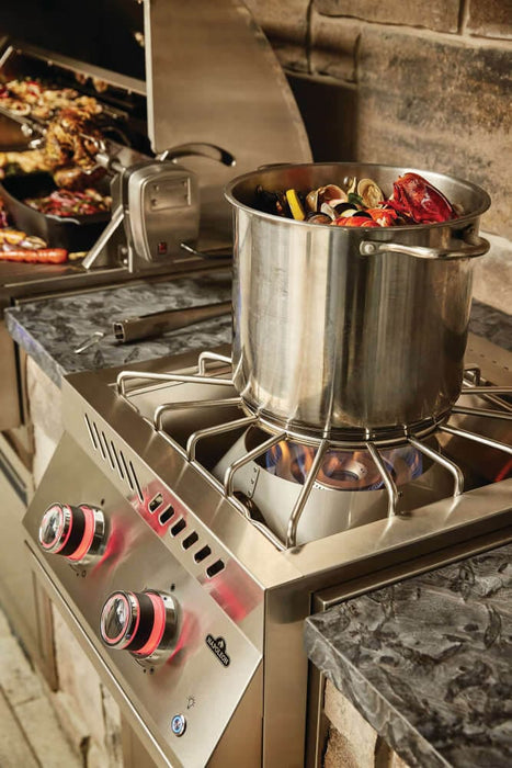 Stainless steel outdoor burner with a pot of vegetables cooking over a blue flame, red-lit control knobs, and stone-accented surroundings.