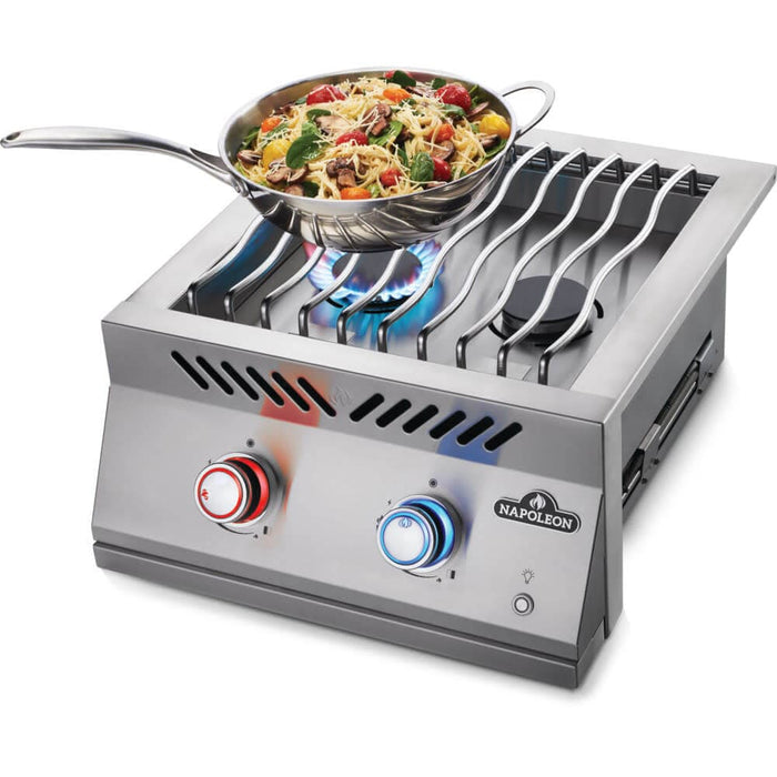 Stainless steel double burner with wavy grill grates, red and blue-lit control knobs, and a skillet of pasta cooking over a blue flame.