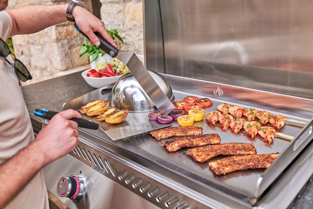 Person grilling various foods, including skewers, ribs, vegetables, and bread, on a stainless steel flat-top griddle in an outdoor kitchen.