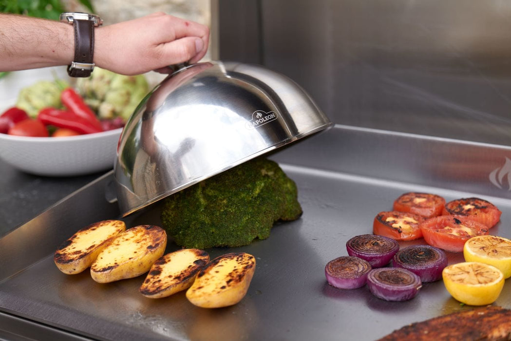 Grilled vegetables, including potatoes, tomatoes, and onions, on a stainless steel griddle, with a hand lifting a domed lid over broccoli.