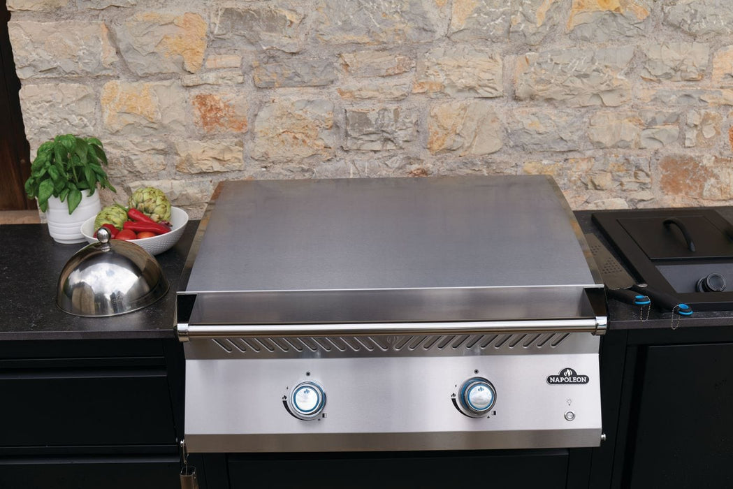 Outdoor stainless steel griddle with a closed lid, blue-lit knobs, surrounded by vegetables and a domed lid, set against a stone wall.