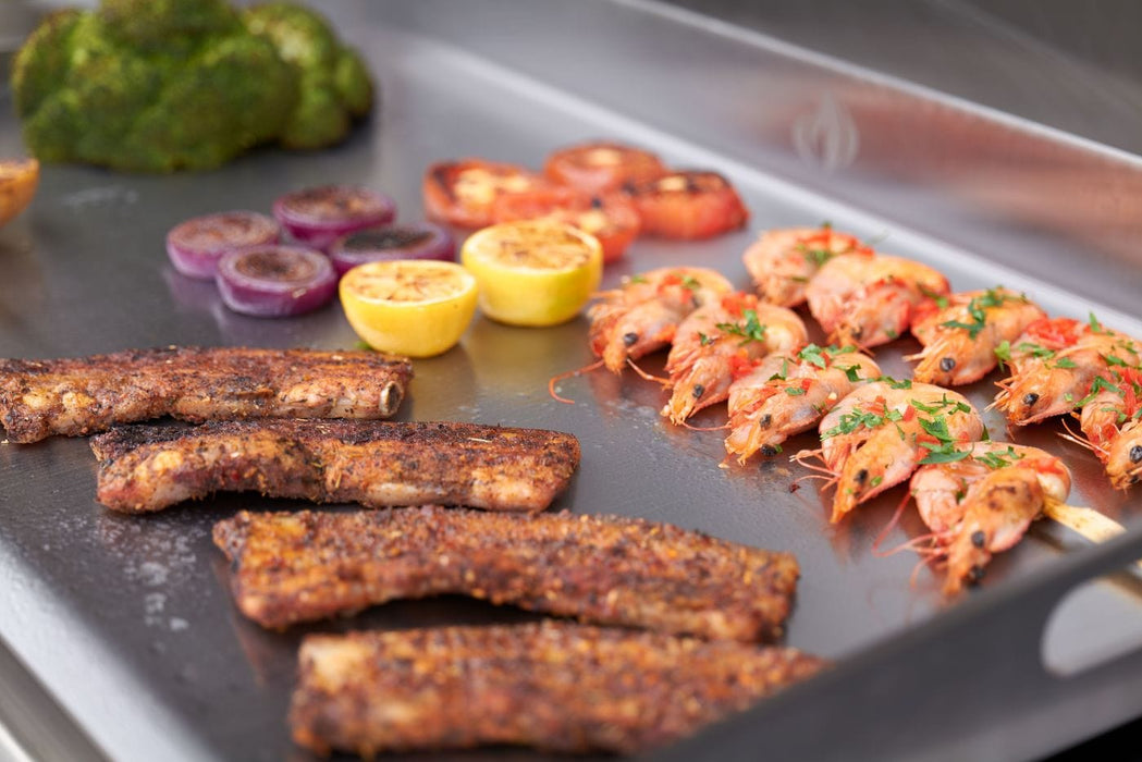 Close-up of grilled food on a stainless steel griddle, featuring shrimp, ribs, broccoli, onions, tomatoes, and lemon halves.