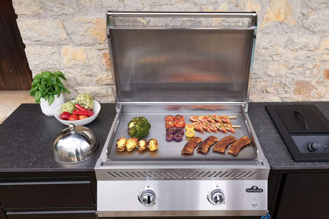 Outdoor stainless steel griddle with an open lid, grilling shrimp, ribs, vegetables, and lemons, surrounded by fresh produce on a countertop.