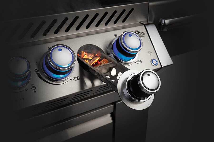 Close-up of a gas grill's control panel with blue-lit knobs and a smoker tube loaded with wood chips, integrated into a sleek design.