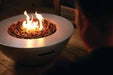 Close-up of a round fire bowl with glowing flames and lava rocks, placed on a wooden deck with a person sitting nearby.